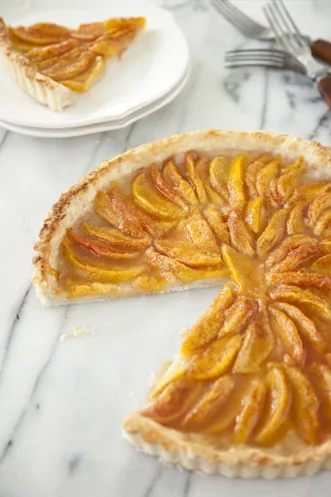 Paula Deen peach tart with fanned glazed peach slices on a golden crust, cut on a marble counter with a slice on a white plate