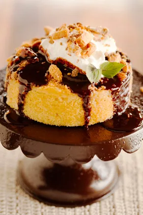 Paula Deen peanut butter chocolate crunch cake slice topped with whipped cream, crushed candy and chocolate drizzle on a cake stand