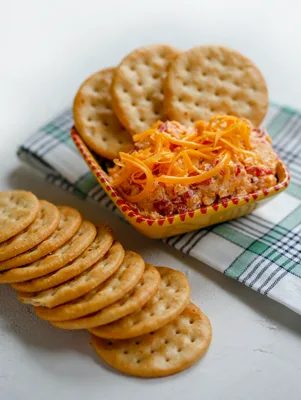 Paula Deen pimento cheese dip in a small red-trimmed dish topped with shredded Cheddar, served with round crackers