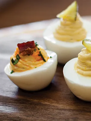 Paula Deen pimiento cheese bacon deviled farm eggs piped with creamy filling and topped with bacon, chives and a lime wedge on a wooden board
