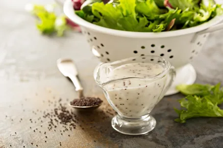 Paula Deen poppy seed dressing in a glass gravy boat with a spoon of poppy seeds and a bowl of fresh salad greens on a stone surface