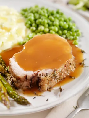 Paula Deen pressure cooker pork loin sliced and covered in brown gravy with green peas, mashed potatoes and asparagus on a white plate