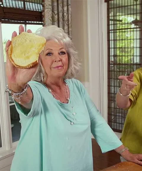 Paula Deen holding a toasted sandwich bun while a woman stirs sloppy joe filling in a skillet in a bright kitchen