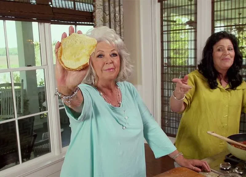 Paula Deen holding a toasted sandwich bun while a woman stirs sloppy joe filling in a skillet in a bright kitchen