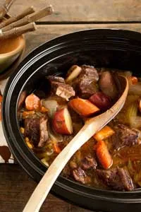 Paula Deen slow cooker beef short ribs with carrots, potatoes and celery in a black slow cooker with a wooden spoon