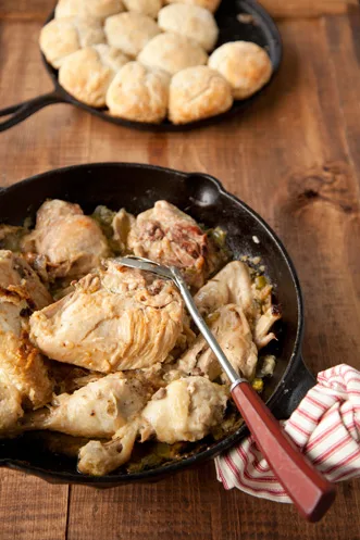 Paula Deen smothered chicken and biscuits with browned chicken pieces in a cast iron skillet and golden biscuits in a second skillet behind it