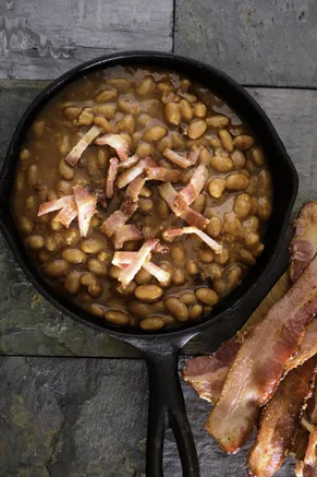 Paula Deen Southern baked beans topped with crumbled bacon in a cast iron skillet with strips of bacon beside it