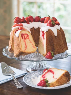 Paula Deen strawberry swirl pound cake on a glass cake stand with white glaze and fresh strawberries, with a pink swirl in the cut slice