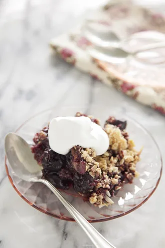 Paula Deen sweet black cherry crisp with oat and almond topping and a dollop of whipped cream on a pink glass plate