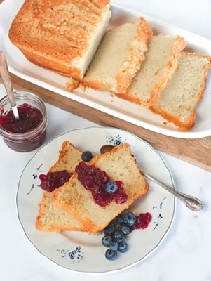Toasted pound cake with blueberry coulis and fresh blueberries on a blue-rimmed plate beside a loaf of pound cake and a jar of jam