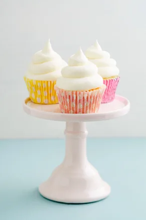 Paula Deen vanilla buttercream frosting piped in tall white swirls on three cupcakes in yellow and pink liners on a pink cake stand